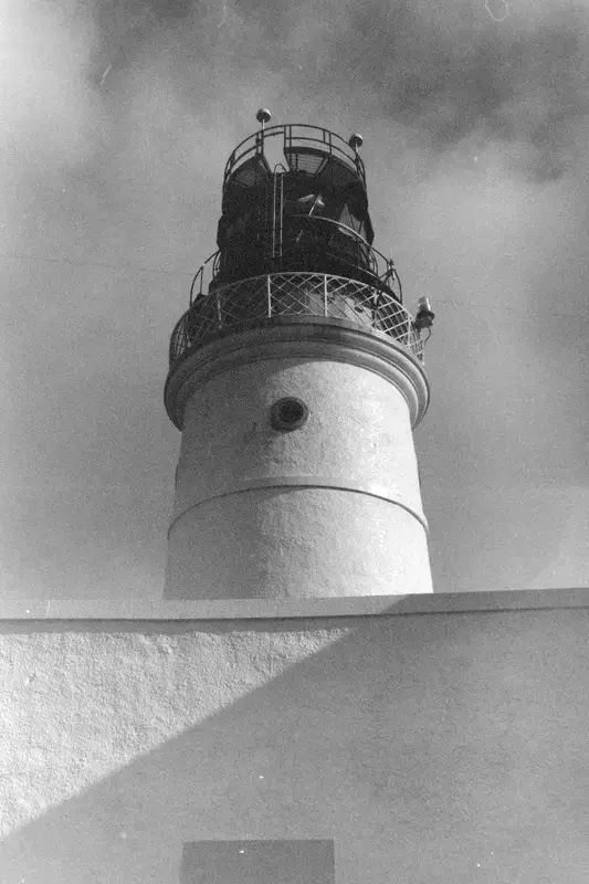 A black-and-white close view of a lighthouse lantern room against a cloudy sky.